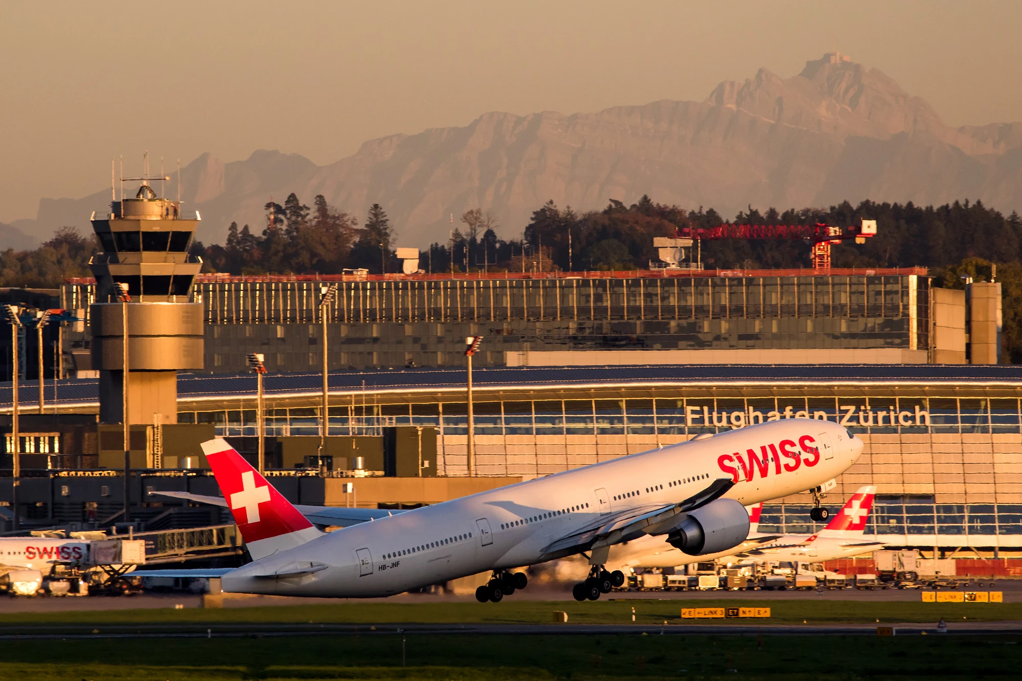 Swiss Boeing 777 rotating off runway 16 at Zurich Airport in evening light, tower and terminal behind, Alps in the background